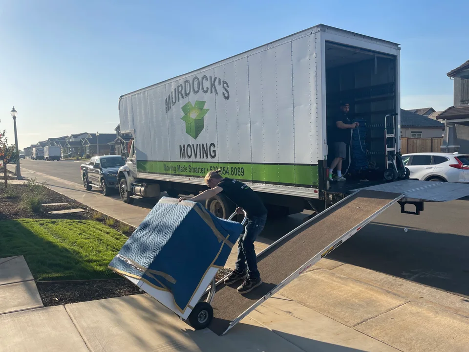 Two Murdock's Moving crew members in branded shirts carry a blue-wrapped appliance past the box truck on a curb