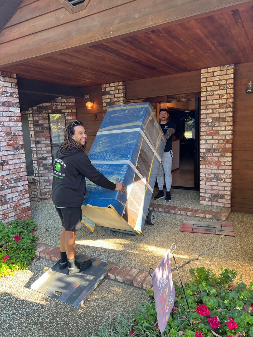 Two smiling Murdock's Moving crew members in branded hoodies carry a blue shrink-wrapped furniture piece up brick front steps
