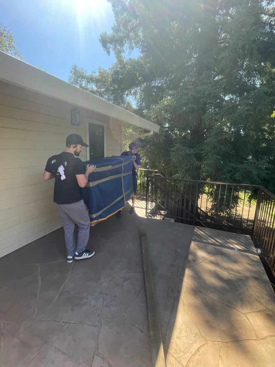 Two Murdock's Moving & Storage movers carry a blanket-wrapped item down stone patio steps using lifting straps