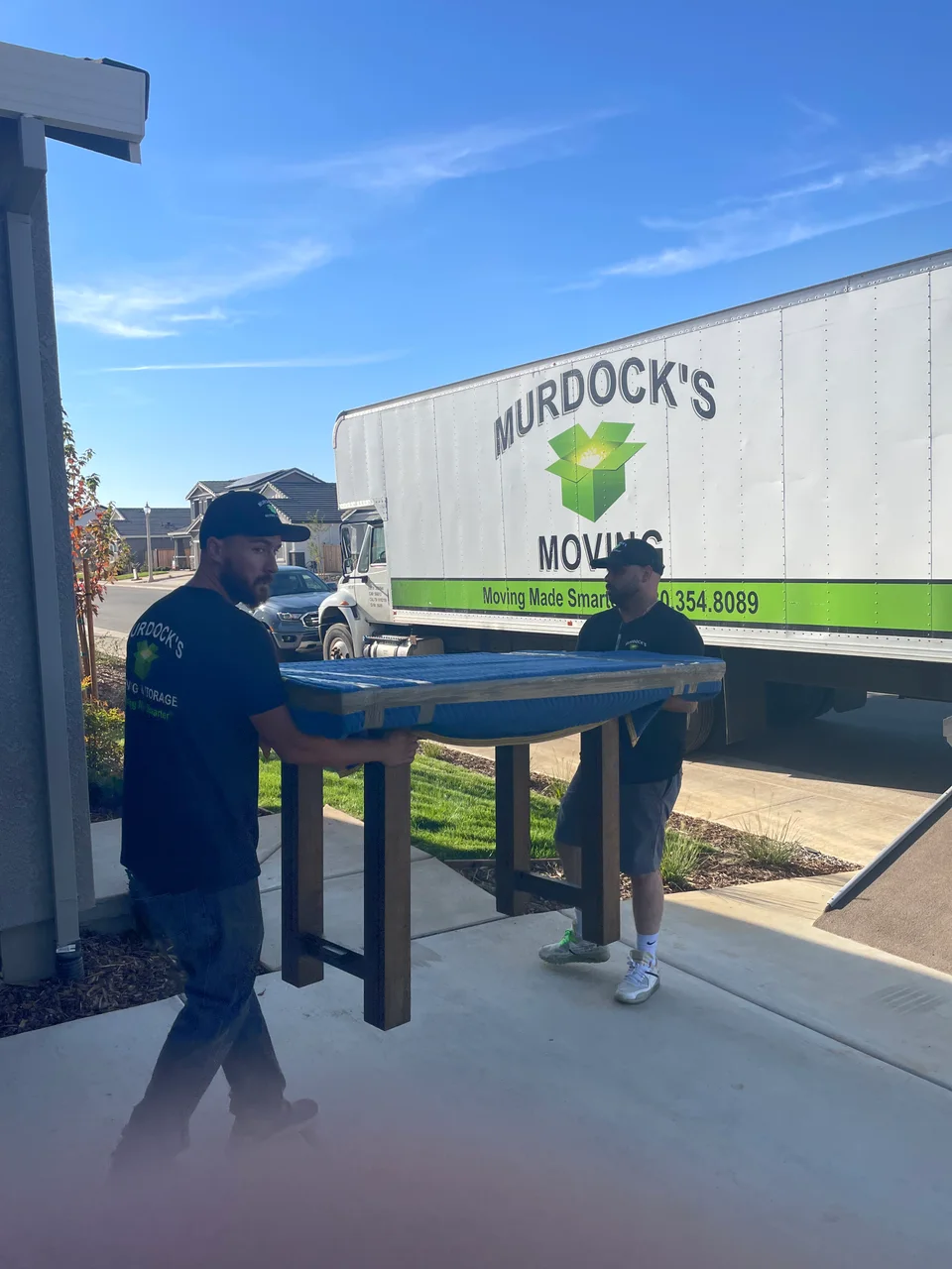 Murdock's Moving crew member guides a blue-wrapped item on a dolly down the truck ramp while a coworker works in the truck