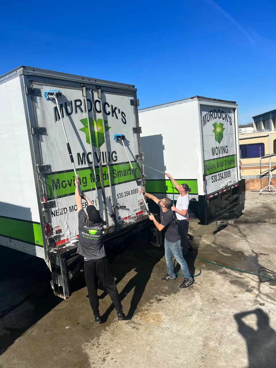 Murdock's Moving & Storage crew washing two branded box trucks under a bright blue sky