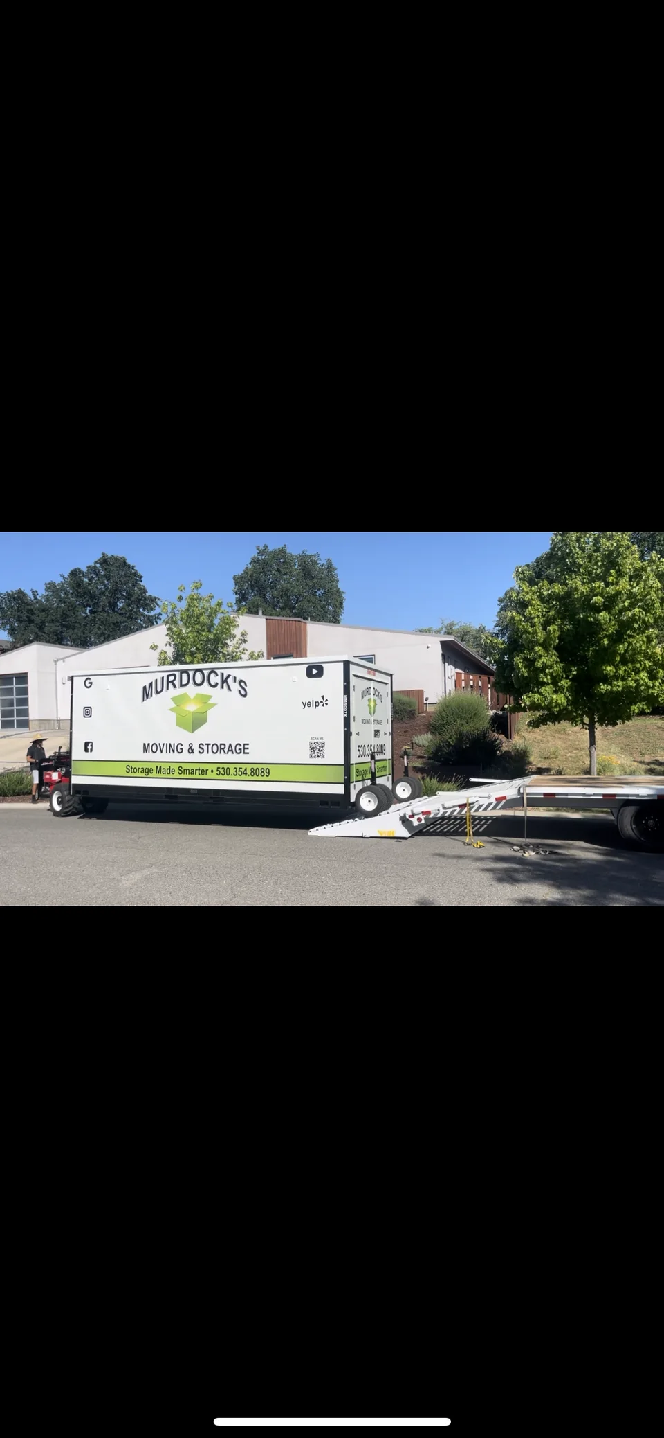 Murdock's Moving & Storage MODS portable storage container beside a flatbed trailer with loading ramp in a driveway