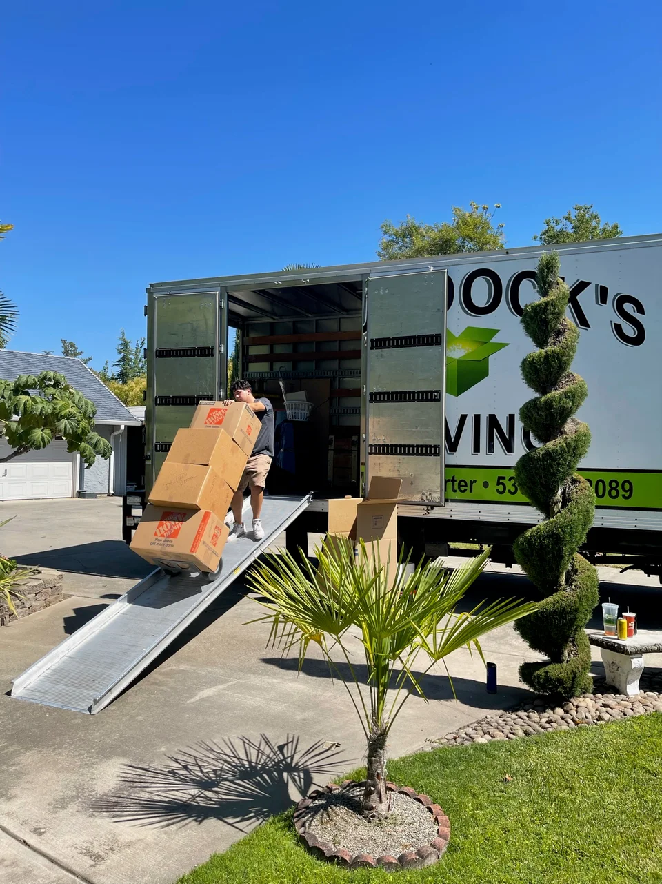 Murdock's Moving & Storage mover wheels a stack of packed boxes down the loading ramp of a branded truck