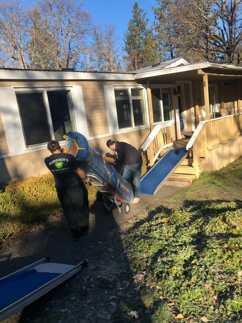 Murdock's Moving & Storage mover guides a hand truck of wrapped furniture up a blue ramp into a home
