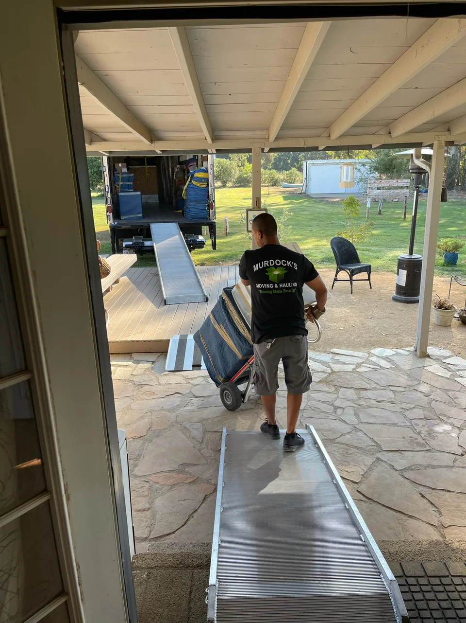 Murdock's Moving & Storage mover guides a hand truck of blanket-wrapped furniture down a ramp from the moving truck