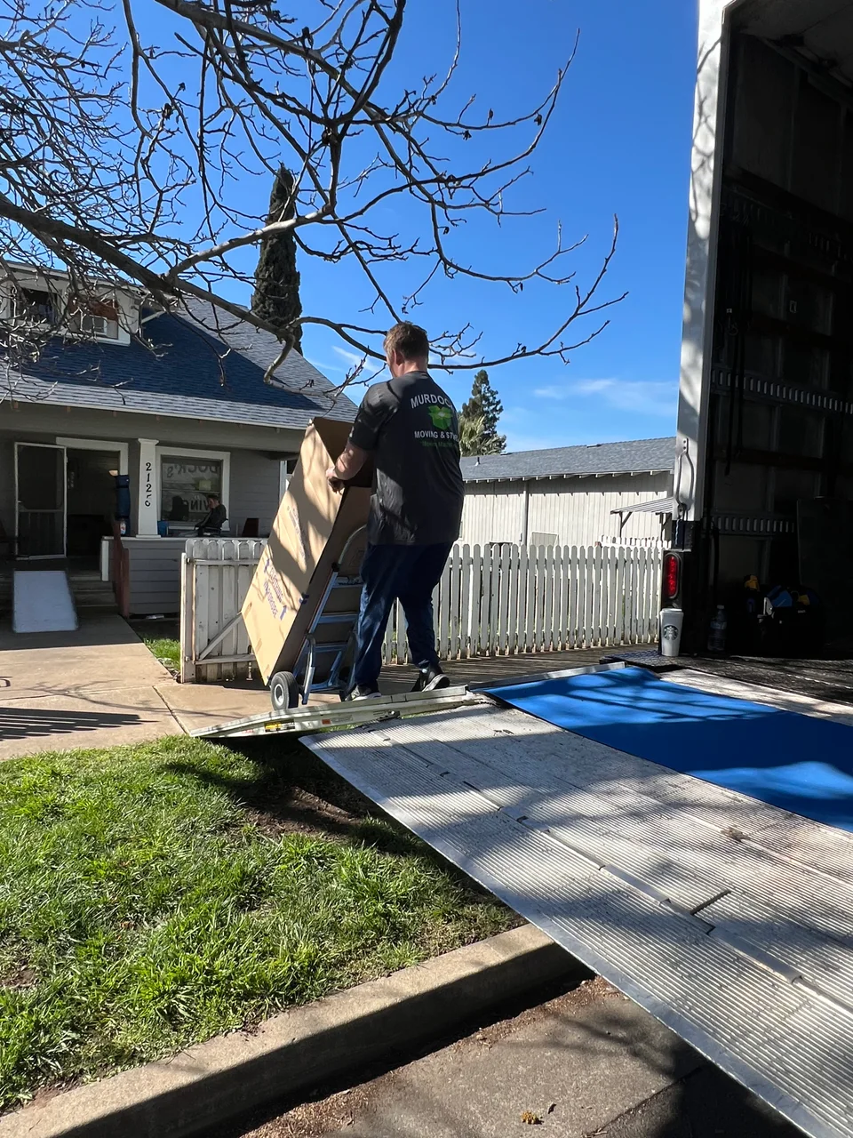 Murdock's Moving & Storage mover rolls a stack of boxes down the ramp of a moving truck outside a home