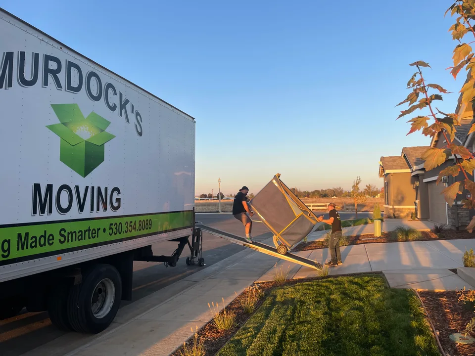 Murdock's Moving crew member rolls a blue-wrapped piece down the box-truck ramp on a dolly while a second mover works inside