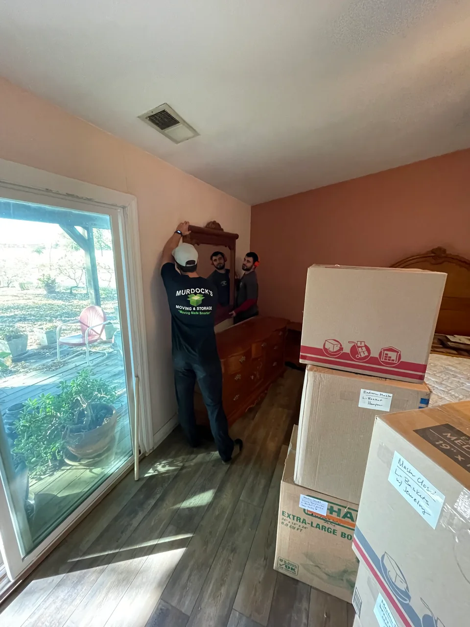 Two Murdock's Moving & Storage crew members carefully carrying a framed mirror past stacked labeled moving boxes indoors