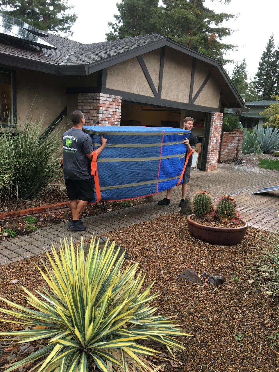 Two Murdock's Moving & Storage movers use orange harness straps to carry a blanket-wrapped item up a home's front walkway