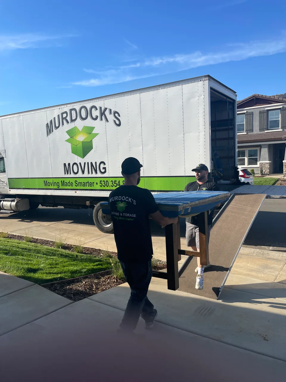 Two Murdock's Moving crew members carry padded, shrink-wrapped table legs and top alongside the branded box truck