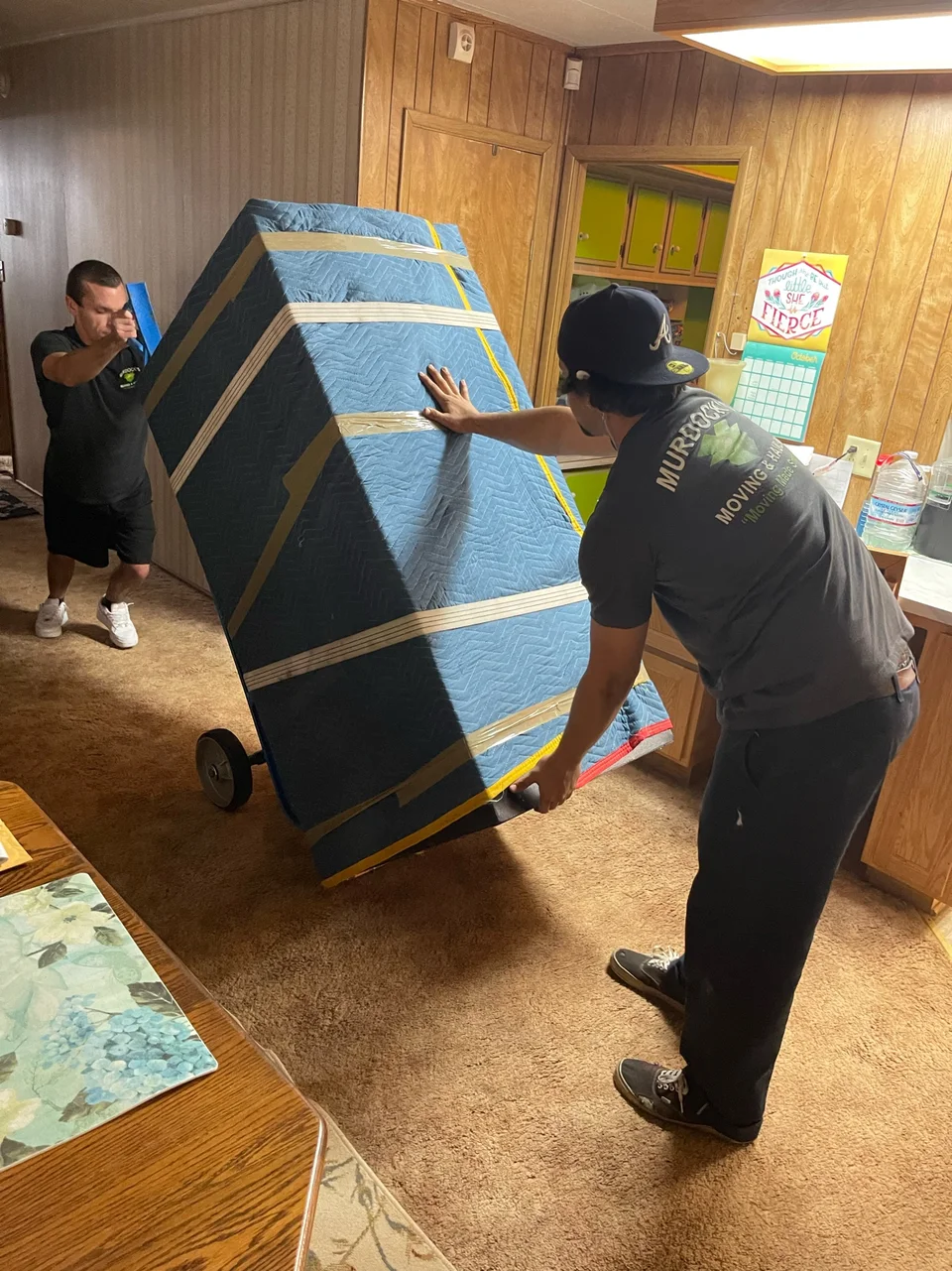 Two Murdock's Moving & Storage movers maneuver a blanket-wrapped mattress on a hand truck inside a home