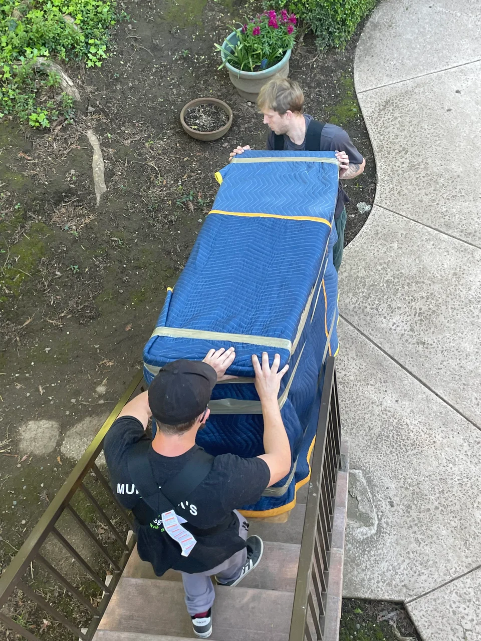Overhead view of two Murdock's Moving & Storage crew members carrying a blanket-wrapped item down garden steps