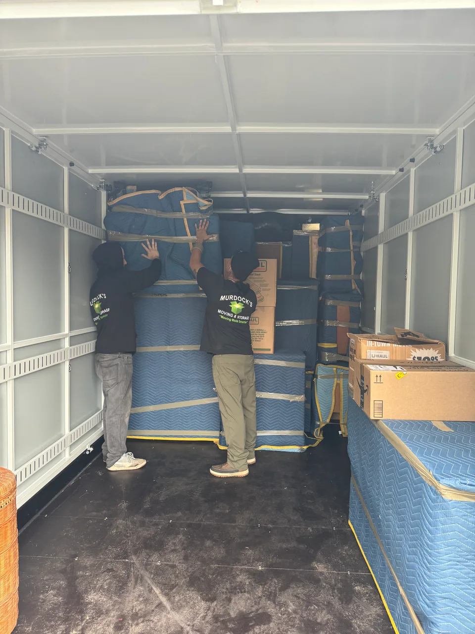 Two Murdock's Moving & Storage movers carefully stack blanket-wrapped furniture inside a loaded moving truck