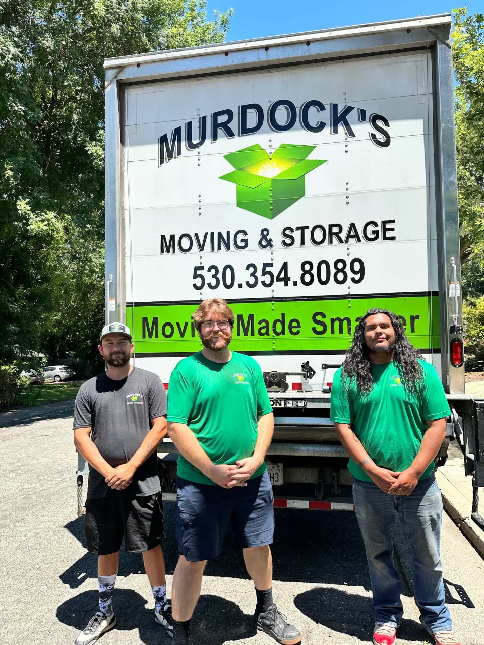 Three smiling Murdock's Moving & Storage movers stand in front of the truck's rear logo and phone number