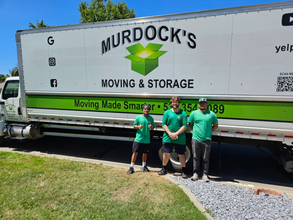 Three Murdock's Moving & Storage crew members in green shirts give thumbs up beside the branded box truck