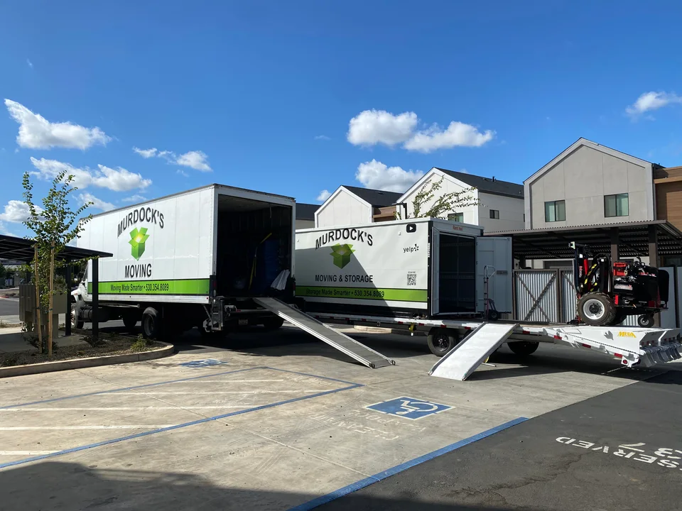 Murdock's Moving box truck beside a branded MOD storage container on a trailer with a loading tractor under blue sky