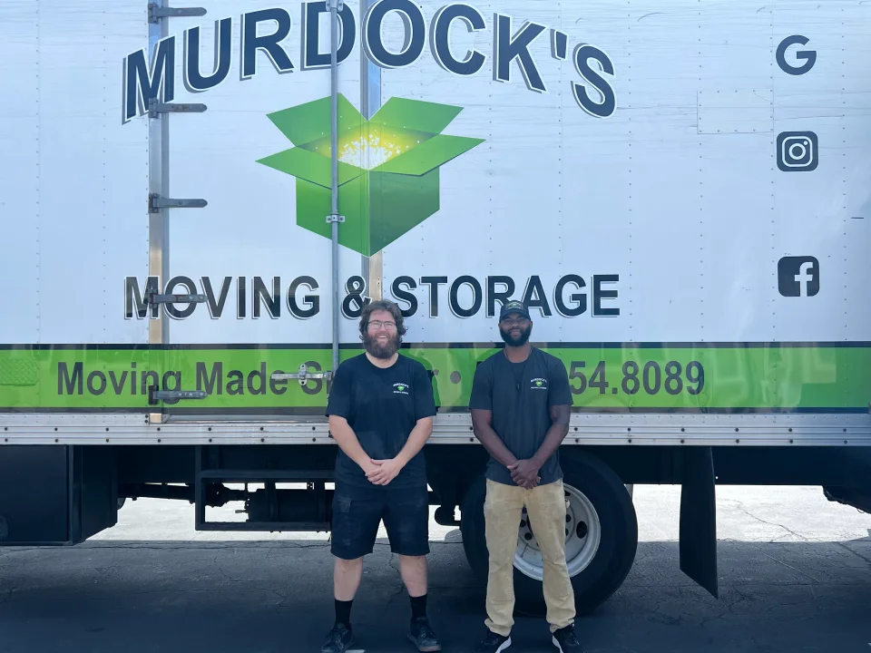 Two smiling Murdock's Moving & Storage crew members standing in front of the large branded truck